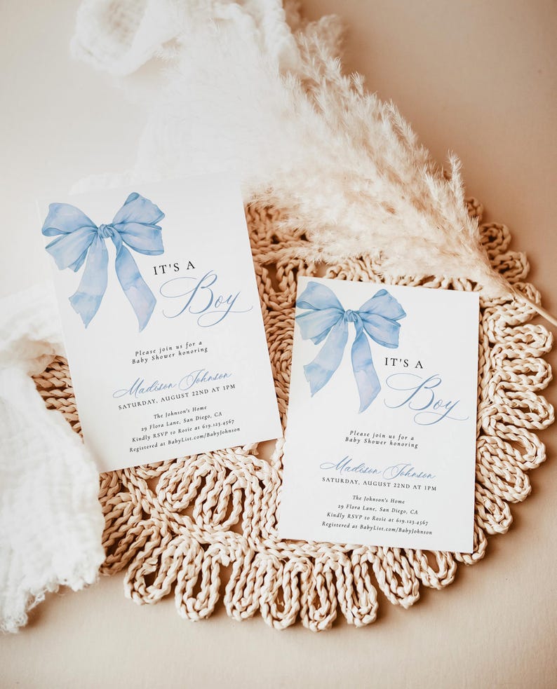 May include: Two blue and white baby shower invitations with a watercolor bow and the text "It's a Boy". The invitations are on a woven placemat.