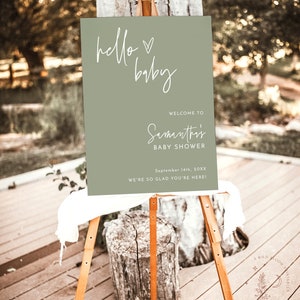 May include: A green welcome sign with white calligraphy that reads "hello baby" and "Samantha's Baby Shower" with the date "September 14th, 20XX". The sign is on an easel and says "We're so glad you're here!"