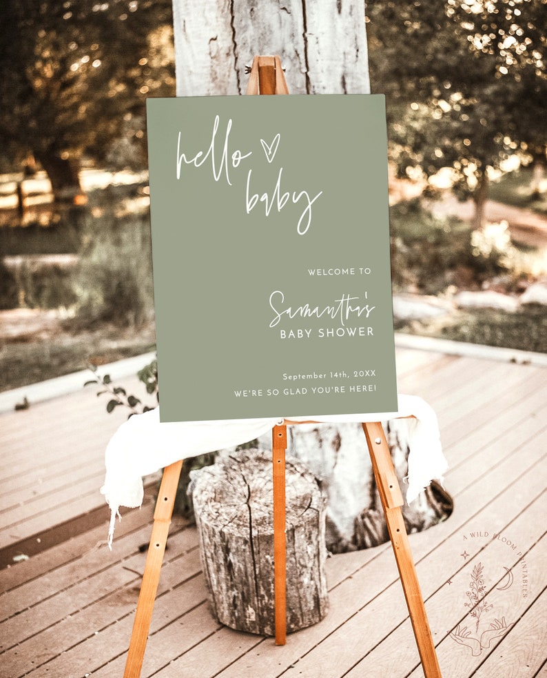 May include: A green welcome sign with white calligraphy that reads "hello baby" and "Samantha's Baby Shower" with the date "September 14th, 20XX". The sign is on an easel and says "We're so glad you're here!"