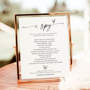 May include: A white sign with a gold frame, featuring the text "i spy" in black cursive font. The sign lists a variety of wedding-themed items to find in a photo booth, such as "bride and groom laughing" and "a funny selfie". The sign also includes the hashtag #HADLEYEVERAFTER.