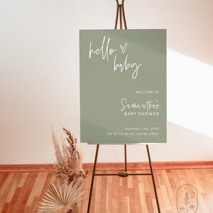 May include: A green welcome sign with white text that reads "hello baby" and "Welcome to Samanthi's Baby Shower" with the date "September 14th, 20XX" and the text "We're so glad you're here!" The sign is on a black easel.