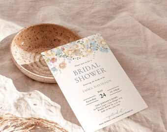May include: A bridal shower invitation with floral accents in pastel colors. The invitation includes the text "BRIDAL SHOWER" and details for the event, including the date, time, and location. A speckled ceramic bowl sits nearby on a white linen cloth.