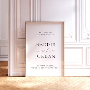 May include: A 24x36 inch wedding welcome sign in a thin wooden frame. The white poster features black text reading: WELCOME TO THE WEDDING OF MADDIE and JORDAN OCTOBER 12, 20XX WE'RE SO GLAD YOU'RE HERE. The design uses a mix of serif and script fonts.