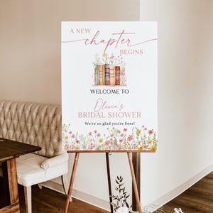 May include: A white foam board sign on a wooden easel. The sign has text that reads "A New Chapter Begins" and "Welcome to Olivia's Bridal Shower". The sign is decorated with floral and book illustrations.