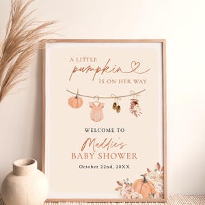 May include: A rectangular framed poster with a cream background featuring the text A LITTLE PUMPKIN IS ON HER WAY, WELCOME TO Maddie's BABY SHOWER, and October 22nd, 20XX. It displays a clothesline with a pumpkin, onesie, shoes, and flowers.