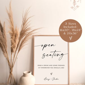 May include: A white sign with black text that reads "open seating" and "Grab a drink and some friends. Sit wherever you would like!" with the names Avery + Jorden at the bottom. The sign is framed in wood and is sitting on a table with a vase and dried pampas grass.