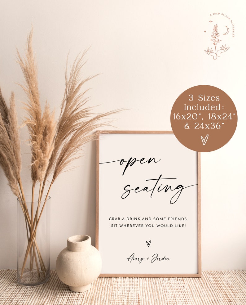 May include: A white sign with black text that reads "open seating" and "Grab a drink and some friends. Sit wherever you would like!" with the names Avery + Jorden at the bottom. The sign is framed in wood and is sitting on a table with a vase and dried pampas grass.