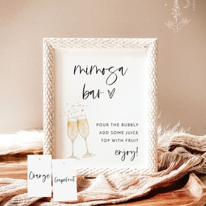 May include: A white framed sign with the text "mimosa bar" in black script. The sign also includes a watercolor illustration of two champagne flutes with gold glitter. Below the illustration, the text reads "POUR THE BUBBLY ADD SOME JUICE TOP WITH FRUIT enjoy!" Two white tags with the text "Orange" and "Grapefruit" are visible in the foreground.