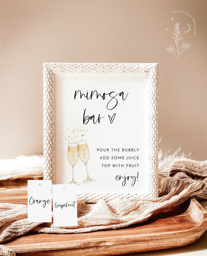 May include: A white framed sign with the text "mimosa bar" in black script. The sign also includes a watercolor illustration of two champagne flutes with gold glitter. Below the illustration, the text reads "POUR THE BUBBLY ADD SOME JUICE TOP WITH FRUIT enjoy!" Two white tags with the text "Orange" and "Grapefruit" are visible in the foreground.