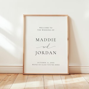 May include: A vertical wedding welcome sign in a light wood frame featuring black text on a white background that reads: WELCOME TO THE WEDDING OF MADDIE and JORDAN OCTOBER 12, 20XX WE'RE SO GLAD YOU'RE HERE. The sign measures 18x24 inches.