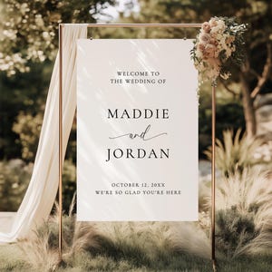 May include: A white rectangular wedding welcome sign on a copper-colored metal stand. The text reads: WELCOME TO THE WEDDING OF MADDIE and JORDAN OCTOBER 12, 20XX WE'RE SO GLAD YOU'RE HERE. A floral arrangement is attached to the top right corner.