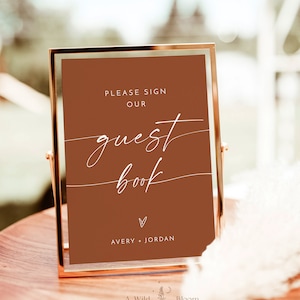 May include: A gold-framed sign with a brown background and white text that reads "Please sign our guest book" with a heart and the names Avery + Jordan. The sign is on a wooden table.