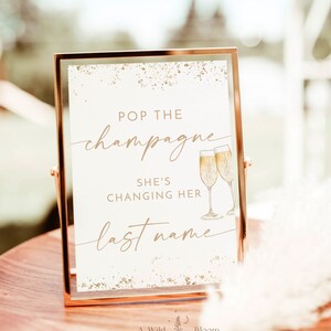 Könnte beinhalten: Ein goldfarbener Rahmen mit einem weißen Hintergrund und goldenem Glitzer. Das Schild lautet "Pop the champagne, she's changing her last name". Zwei Champagnergläser sind neben dem Text abgebildet.