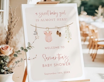 Op de afbeelding: Een rechthoekig 46x61 cm schuimrubberen bord op een houten ezel. Het bord bevat de tekst A sweet baby girl IS ALMOST HERE! WELCOME TO Samantha's BABY SHOWER September 14th, 20XX, met een illustratie van een waslijn met babyartikelen.