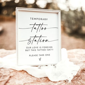 以下が含まれることがあります： 「Temporary tattoo station. Our love is forever but this tattoo isn't! Please take one.」と黒文字で書かれた白い看板。看板は白い布と茶色の岩の上に置かれている。