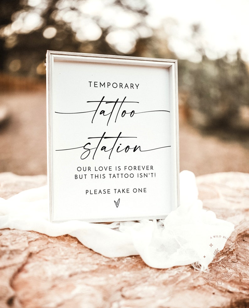 以下が含まれることがあります： 「Temporary tattoo station. Our love is forever but this tattoo isn't! Please take one.」と黒文字で書かれた白い看板。看板は白い布と茶色の岩の上に置かれている。