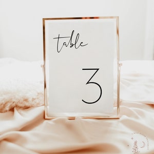 May include: A white table number card with a gold frame. The card says "table" and the number "3" in a modern font.
