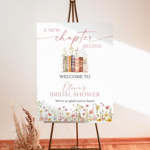 May include: A white foam board sign on a brown easel. The sign reads "A New Chapter Begins" and "Welcome to Olivia's Bridal Shower." Floral illustrations and text are in pink and brown. The sign is 18x24 inches.
