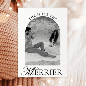 Op de afbeelding: Een wenskaart met een zwart-witfoto van een stel op het strand, omlijst door een boog. De tekst "THE MORE THE" staat boven de foto en "MERRIER" eronder in elegant schrift. De kaart is geplaatst tegen een gezellige, getextureerde achtergrond.