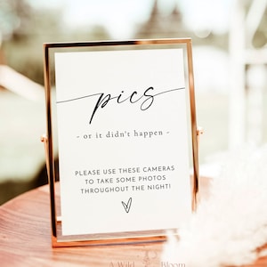 May include: A gold-framed sign with the text "pics - or it didn't happen -" and a message encouraging guests to use the cameras provided to take photos throughout the night. The sign is on a wooden table with a floral design at the bottom.