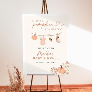 May include: A rectangular 18x24 inch foam board sign featuring the text A LITTLE pumpkin IS ON HER WAY, WELCOME TO Maddie's BABY SHOWER, and October 22nd, 20XX. The design includes illustrations of a pumpkin, baby clothes, and flowers.