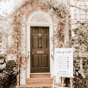 May include: A white sign with black text and icons listing the order of events for a wedding. The sign reads "order of events" and lists the ceremony, cocktail hour, dinner, speeches, cake cutting, first dance, and send off. The sign is on a wooden easel in front of a white door with a black door.