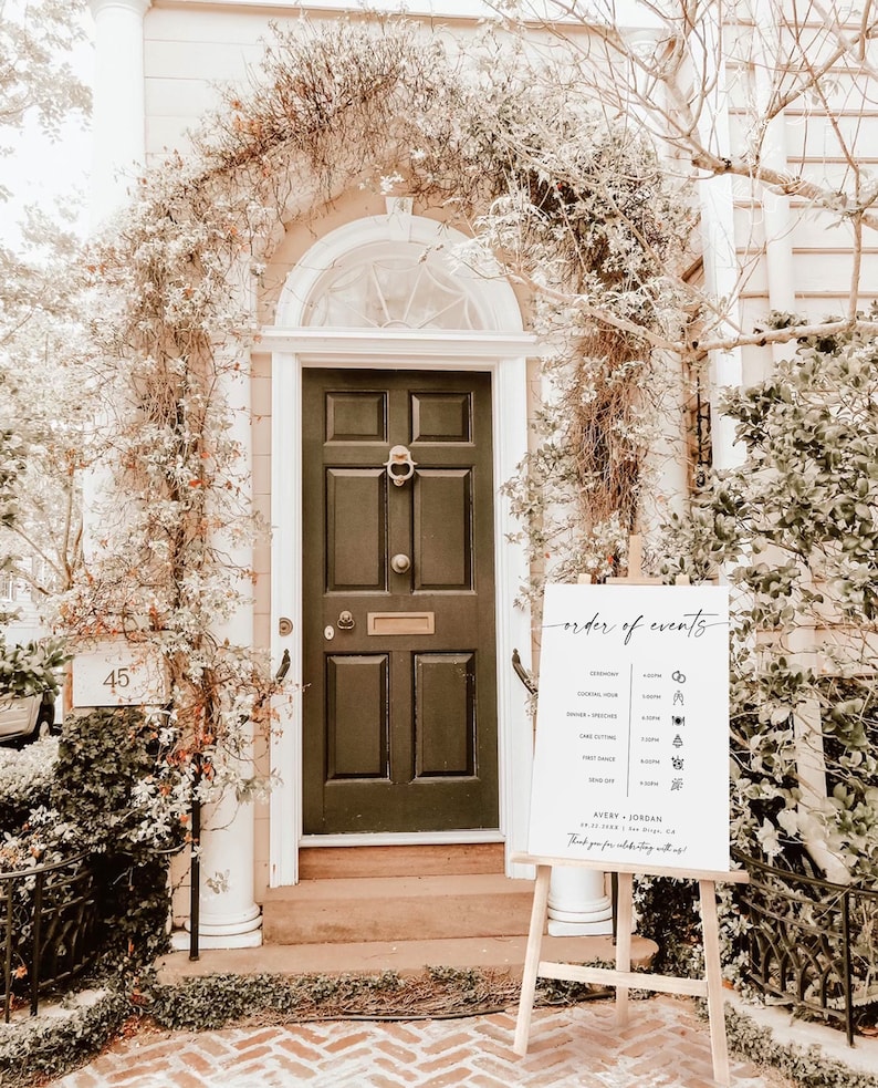 May include: A white sign with black text and icons listing the order of events for a wedding. The sign reads "order of events" and lists the ceremony, cocktail hour, dinner, speeches, cake cutting, first dance, and send off. The sign is on a wooden easel in front of a white door with a black door.
