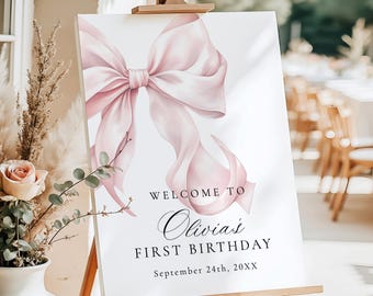 Peut inclure: Un panneau blanc sur un chevalet en bois avec une grande illustration d'un nœud rose. Le panneau indique "BIENVENUE À Olivia's PREMIER ANNIVERSAIRE" avec la date "24 septembre 20XX". Un arrangement floral dans un vase blanc est visible.
