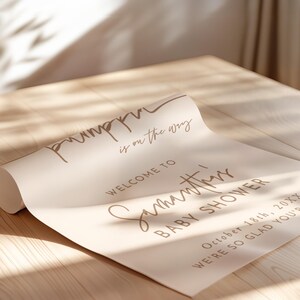 May include: A rolled poster paper sign with a light beige background and brown text that reads: pumpkin is on the way, WELCOME TO Samantha's BABY SHOWER, October 18th, 20XX, WE'RE SO GLAD YOU'RE HERE.