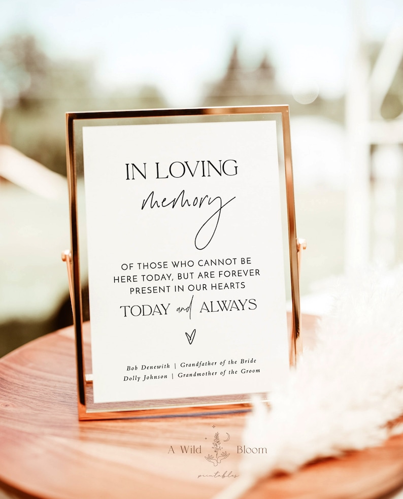 May include: A white sign in a gold frame with the text "In loving memory" and a handwritten script of "memory". The sign also includes the text "Of those who cannot be here today, but are forever present in our hearts today and always". The sign also includes the names "Bob Denewith" and "Dolly Johnson" with their respective titles "Grandfather of the Bride" and "Grandmother of the Groom".