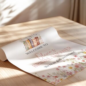 May include: A rolled-up poster with the text "Welcome to Olivia's Bridal Shower" in pink script. The poster features a watercolor illustration of books and wildflowers. The words "We're so glad you're here!" are also included.