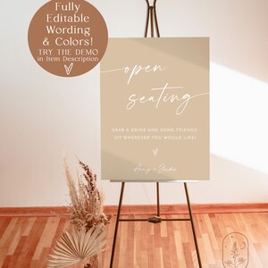 May include: A printable sign with the text "open seating" in white script font on a beige background. The sign also includes the text "Grab a drink and some friends. Sit wherever you would like!" and the names "Aury + Jorden" in white script font. The sign is on a black easel.
