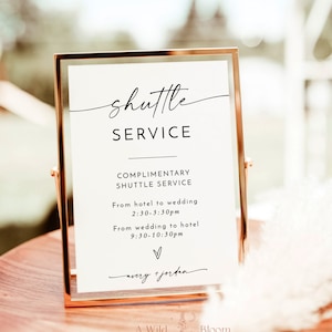 May include: A gold-framed sign with white text that reads "shuttle service" and provides complimentary shuttle service times from the hotel to the wedding and from the wedding to the hotel. The sign also includes the names "avery + jordan" and the logo "A Wild Bloom Designs".