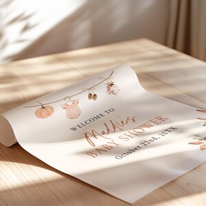 May include: A white poster paper sign featuring a watercolor illustration of a string with a pumpkin, baby clothes, and shoes. The text reads: WELCOME TO Maddies BABY SHOWER October 22nd, 20XX. The design includes autumn-themed foliage accents.