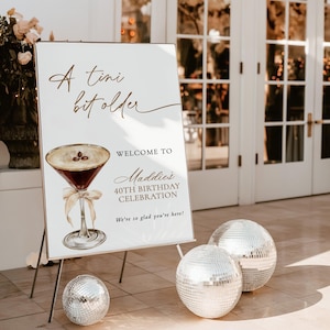 May include: A white foam board sign with the text "A tini bit older" and "Welcome to Maddie's 40th Birthday Celebration". The sign features a cocktail illustration and is propped on a metal stand. Three disco balls are on the floor.