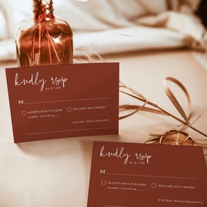 May include: Two brown RSVP cards with white text. The text on the cards reads "kindly rsvp by 8.1.XX". Each card has a section for the guest to indicate whether they will accept or decline the invitation, and a space for the guest to write the number of people attending. There is also a space for the guest to write any dietary requirements.
