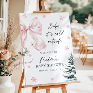 Op de afbeelding: Een wit babyborrelbord op een houten ezel. Het bord heeft een grote roze strik, sneeuwvlokken en de tekst "Baby, it's cold outside". Het bevat ook de tekst "Welcome to Maddie's Baby Shower" en de datum.