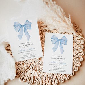Może przedstawiać: Dwa zaproszenia na baby shower z akwarelowym wzorem niebieskiej kokardy. Tekst brzmi "Please join us for a BABY SHOWER honoring Madison Johnson, Saturday, August 22nd at 1PM". Zaproszenia leżą na beżowej, tkanej podkładce.