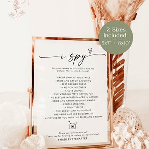 May include: A gold-framed printable sign with the text "i spy" and a list of items to find in a wedding photo booth. The sign is on a light brown background with a white and brown dried flower arrangement. The sign is available in two sizes: 5x7 inches and 8x10 inches.