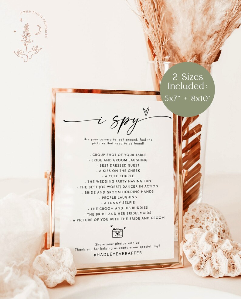 May include: A gold-framed printable sign with the text "i spy" and a list of items to find in a wedding photo booth. The sign is on a light brown background with a white and brown dried flower arrangement. The sign is available in two sizes: 5x7 inches and 8x10 inches.