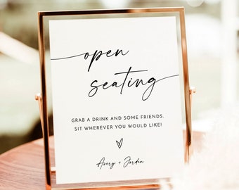 May include: A white sign with black text in a gold frame that reads "open seating" and "Grab a drink and some friends. Sit wherever you would like!" with a heart and the names "Avery + Jorden" below. The sign is on a wooden table.