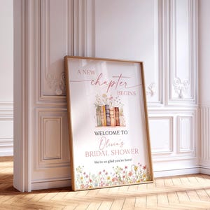 May include: A framed poster with the text "A New Chapter Begins" and "Welcome to Olivia's Bridal Shower". The poster features a floral and book design with a wooden frame. The poster is 24x36 inches.