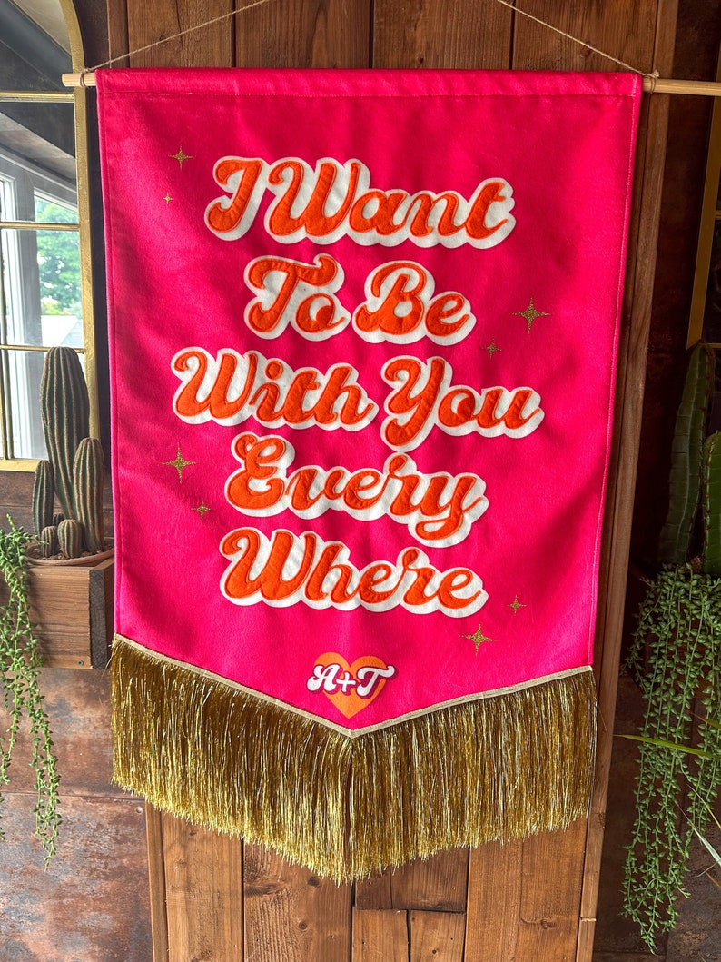 May include: A pink fabric banner with gold fringe and the text "I Want To Be With You Every Where" in white and orange lettering. A heart with the letters "A+T" is in the center of the banner.