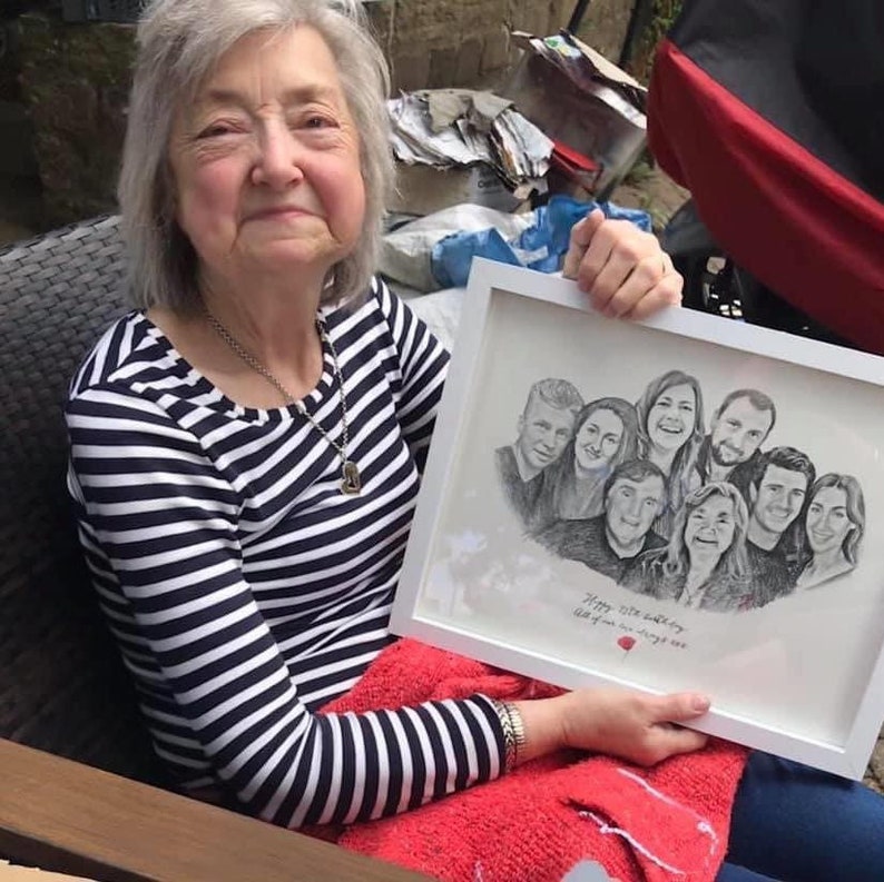 Pu&ograve; includere: Una donna tiene in mano un disegno incorniciato di un gruppo di persone. Il disegno &egrave; in bianco e nero e il testo sottostante recita "Happy 50th birthday to all of you we love you always".
