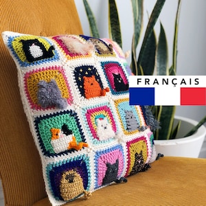 May include: A colorful crocheted pillow with a variety of cat designs. The pillow is made with different colored squares and features a variety of cat faces, including a black cat, a white cat, a gray cat, and a brown cat. The pillow is sitting on a yellow chair.