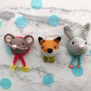 May include: Three crocheted animal head ornaments with colourful scarves. A grey mouse, an orange fox, and a grey rabbit.