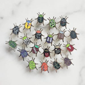 Könnte beinhalten: Eine Sammlung von 20 bunten Käfer-Pins mit verschiedenen Zitaten aus beliebten Büchern und Filmen. Die Pins sind aus Metall und haben eine glänzende Oberfläche.