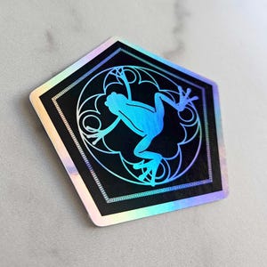 May include: A black and blue holographic sticker with a geometric design featuring a frog silhouette.