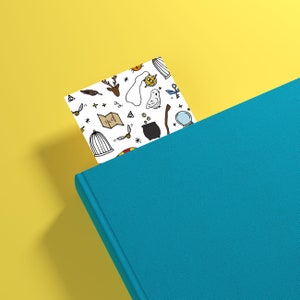 K&ouml;nnte beinhalten: Ein wei&szlig;es Lesezeichen mit einem blauen Buch. Das Lesezeichen zeigt ein Muster aus verschiedenen Harry-Potter-Motiven, darunter eine Eule, ein K&auml;fig, ein Zauberstab, eine Karte und ein Kessel.