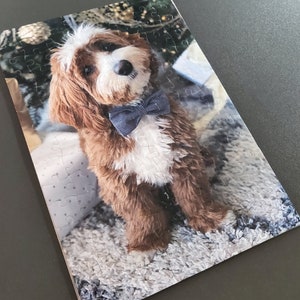 May include: A completed jigsaw puzzle featuring a brown and white dog wearing a blue bow tie. The dog is sitting on a fluffy, light-colored rug in front of a Christmas tree. The puzzle is rectangular and has many pieces.